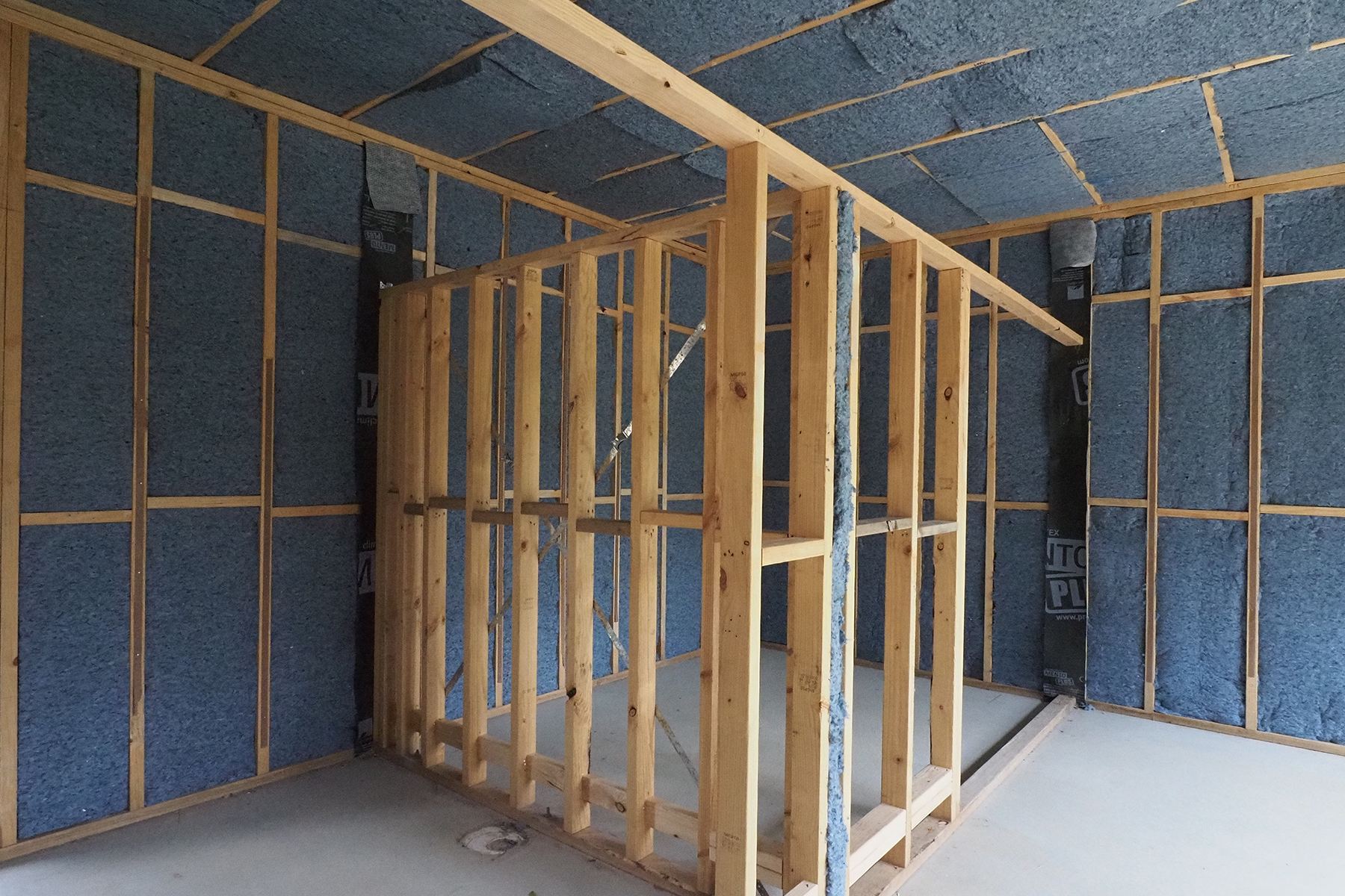 A wide angle view of the corner of a house under construction, the framework is in and the floor is bare concrete. Between the framework the insulation is partially installed. The insulation is a denim blue colour