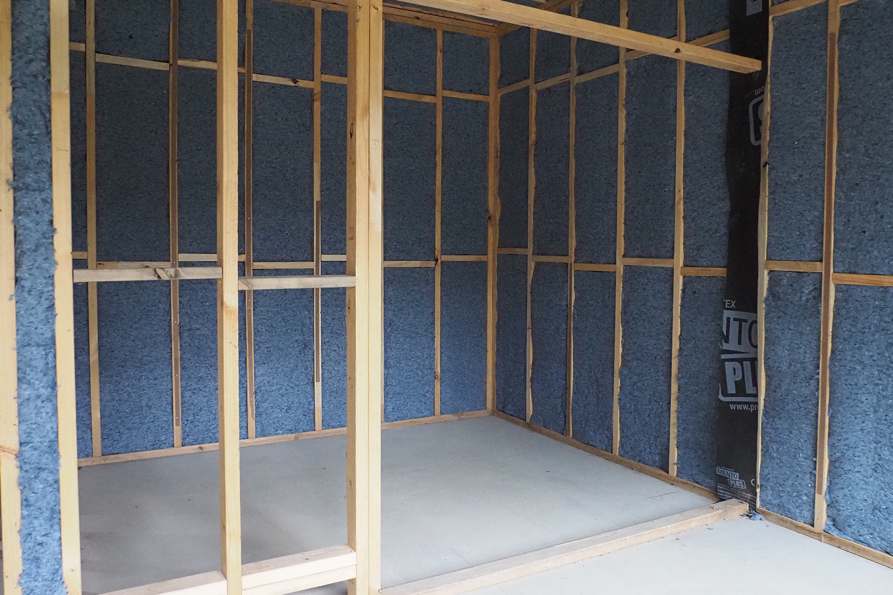 A room in a house under construction, the framework is in and the floor is bare concrete. Between the framework the insulation is partially installed. The insulation is a denim blue colour