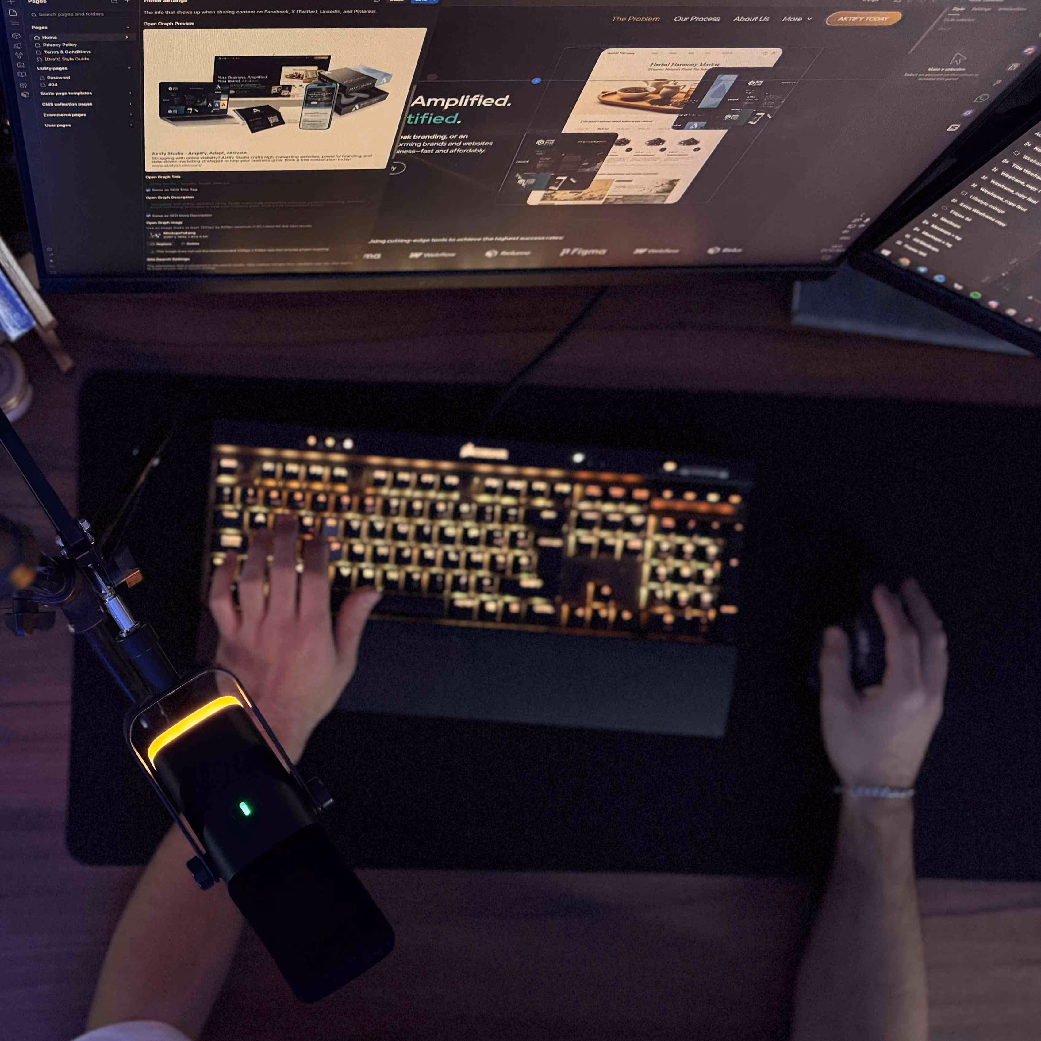 Overhead view of person typing on a backlit keyboard and using a mouse, with a microphone and two computer monitors displaying design software.