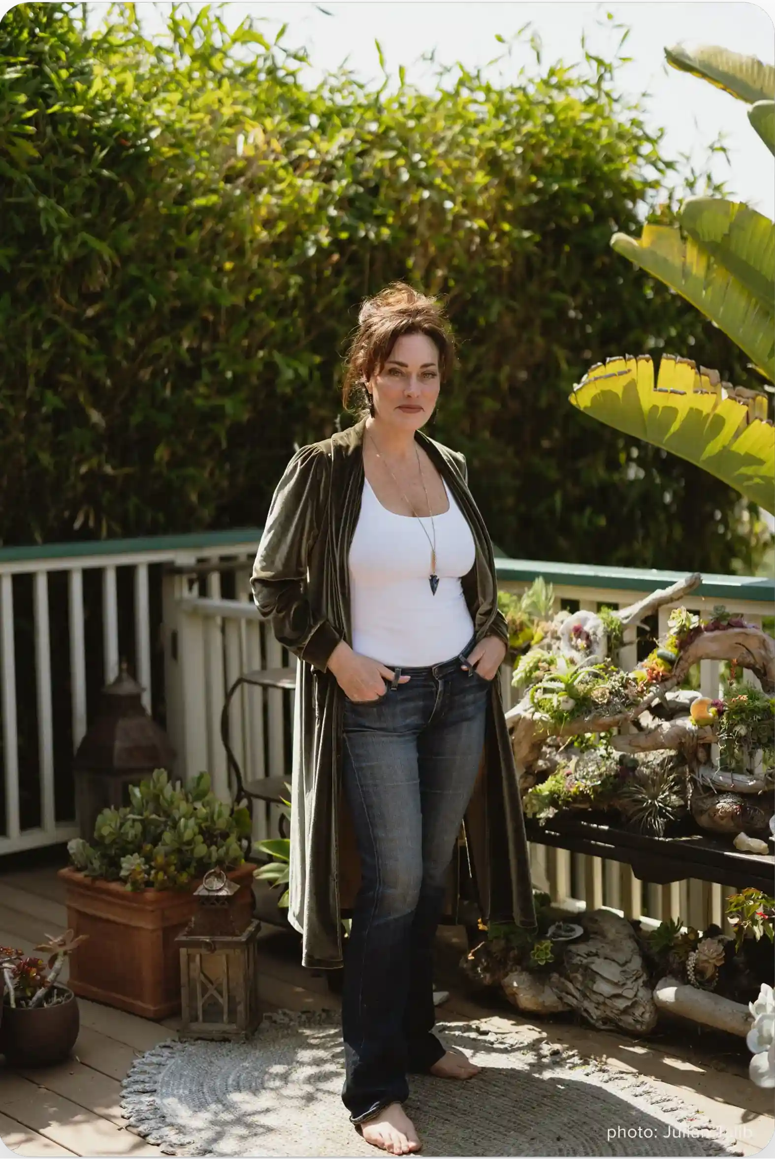 Jennifer Love is standing on a porch, wearing a long green jacket, a white top, and jeans.