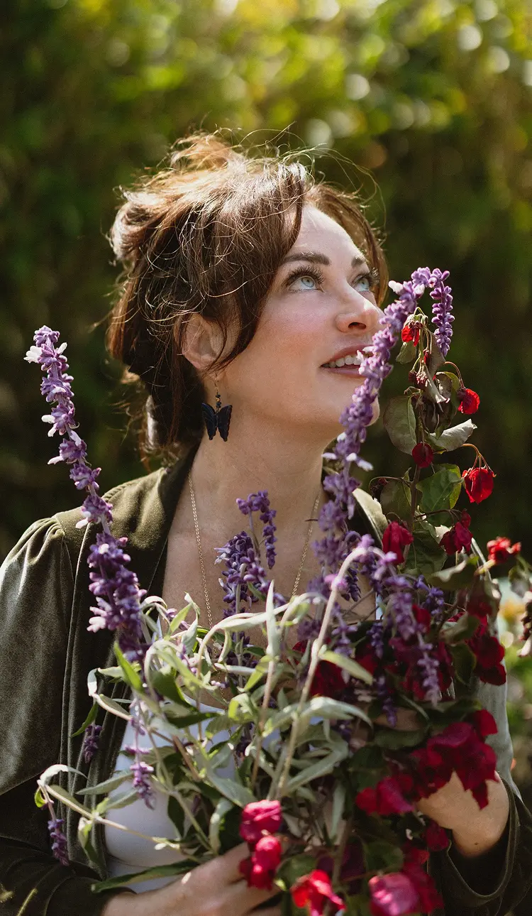 Jennifer Love, JLove. A woman  with dark hair looks upwards while holding a bouquet of purple and red flowers, resembling lavender.