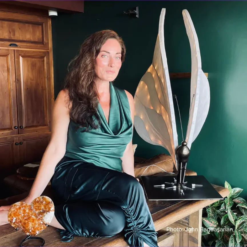 Jennifer Love,  is pictured with long brown hair, wearing a green top and black pants, seated next to a unique, illuminated sculpture