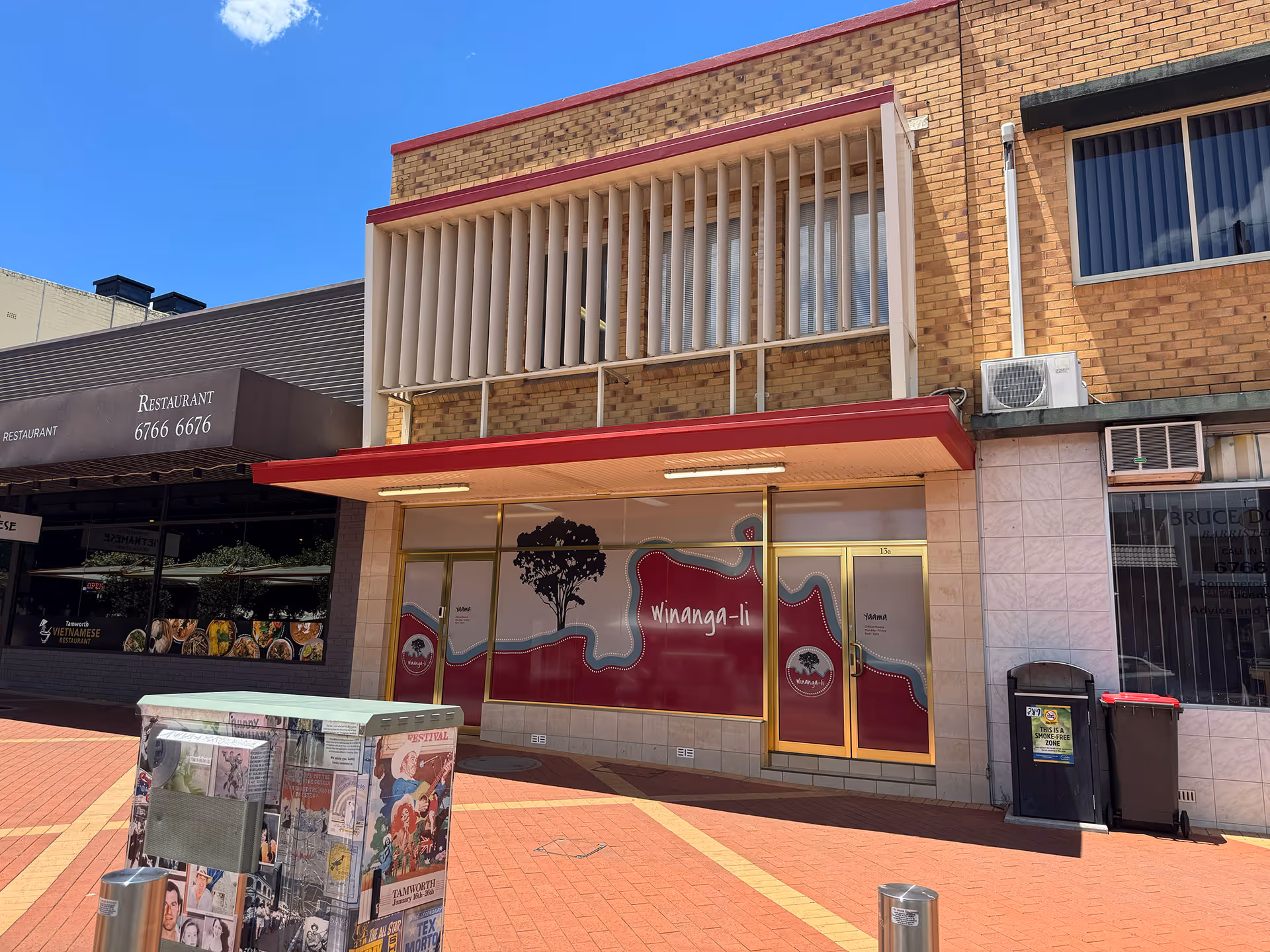 Street view of a brick building with a shop named Winanga-li featuring a red and white storefront with gold-framed windows and door.