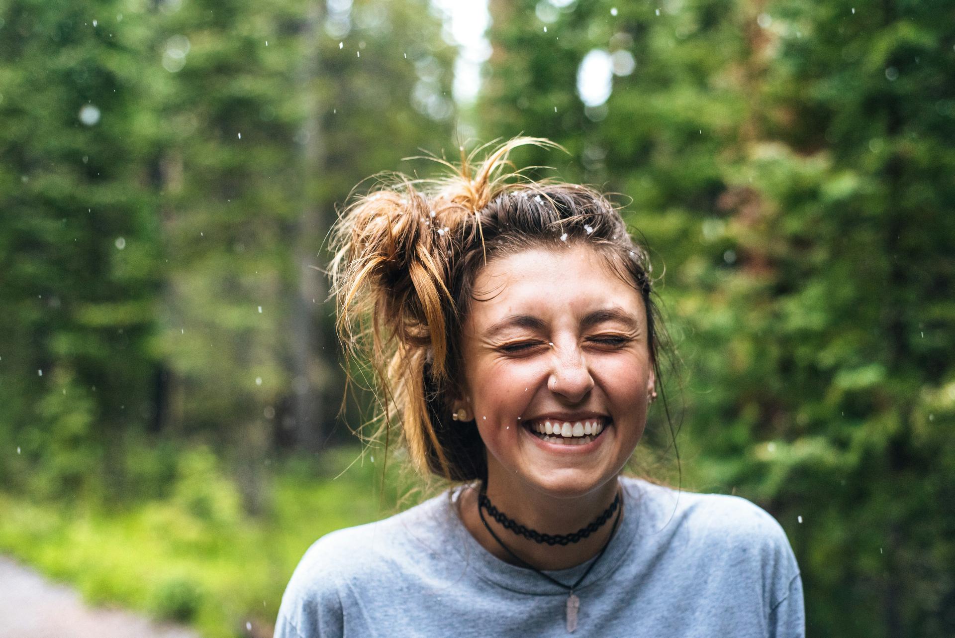 Young woman with a messy bun smiling with eyes closed in a green forest while snow falls.
