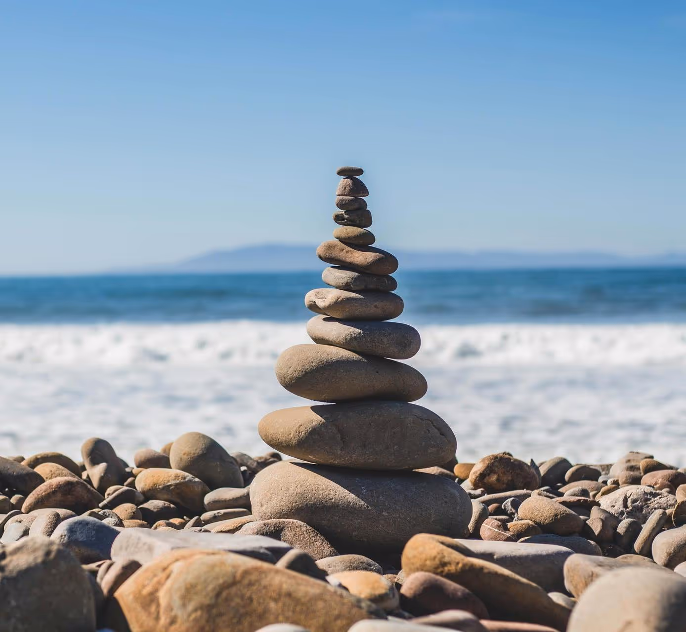 Stack of smooth stones balanced on a rocky beach with ocean waves and a clear blue sky in the background.