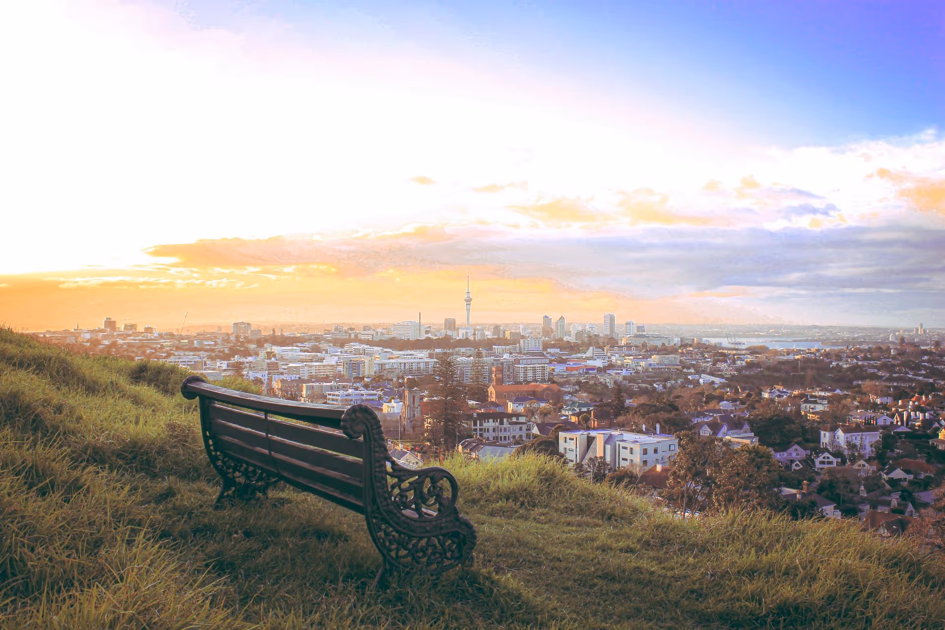 Empty ornate bench on grassy hill overlooking a cityscape under a colorful sunset sky.