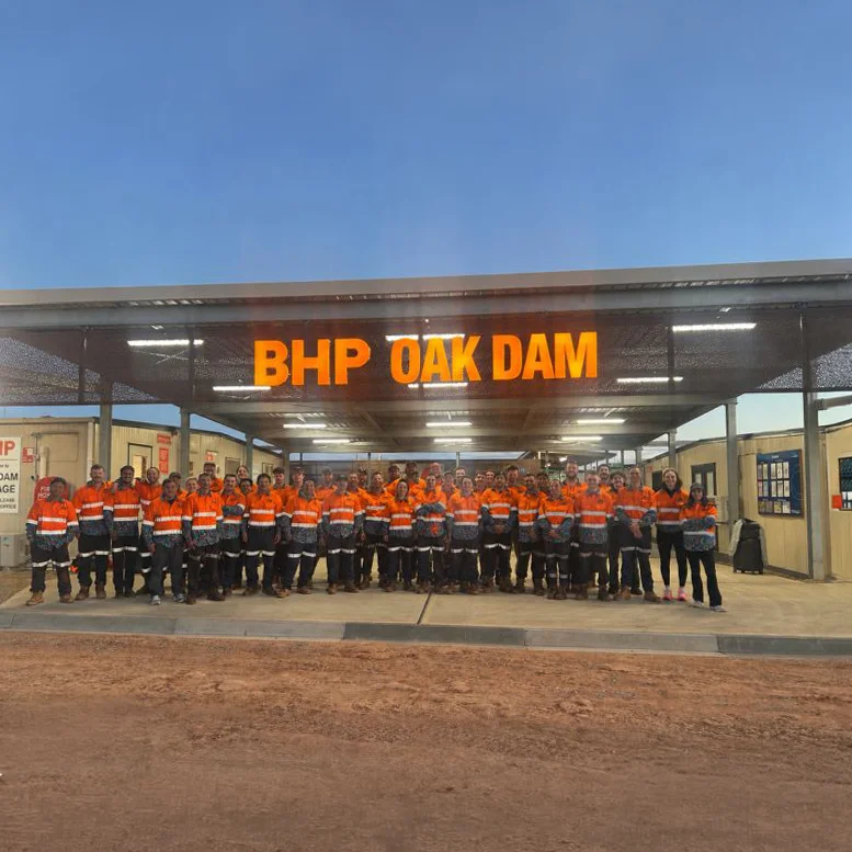 A large group of Titeline workers in orange high-visibility uniforms standing under a canopy with BHP OAK DAM sign.