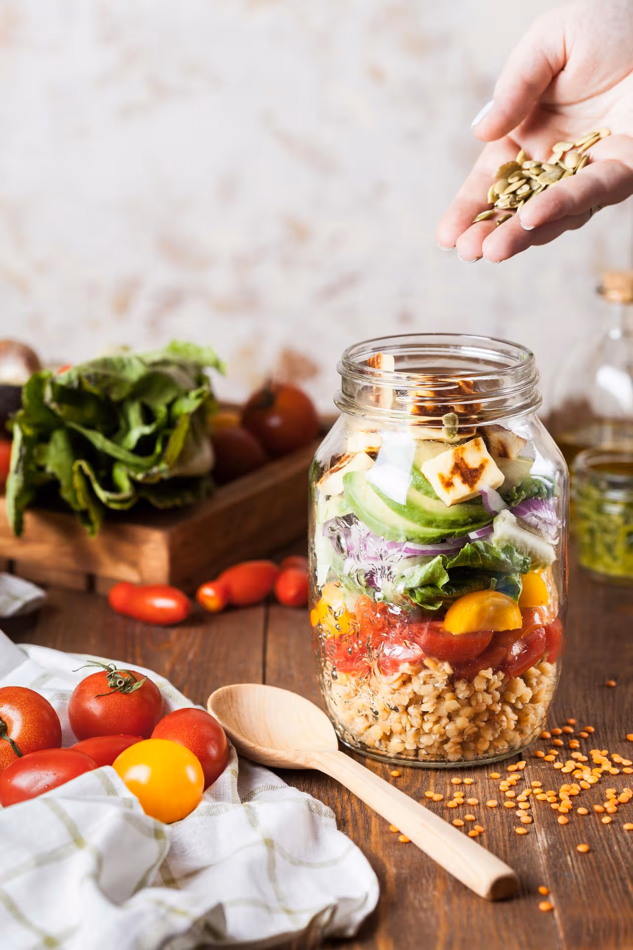 A hand sprinkles seeds over a vibrant salad jar with layers of grains, tomatoes, and greens. Fresh vegetables and a wooden spoon surround the jar.