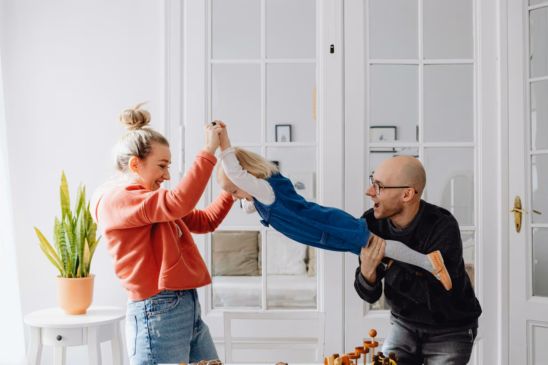 A joyful family moment: A woman and man lift a smiling child in a blue dress, playing indoors near a white wall with glass doors and a green potted plant.