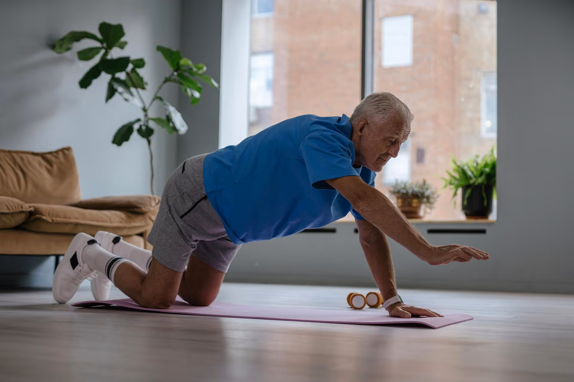An elderly man in a blue shirt and gray shorts exercises on a yoga mat, extending an arm forward. The room has a sofa, potted plants, and large windows, conveying a calm atmosphere.