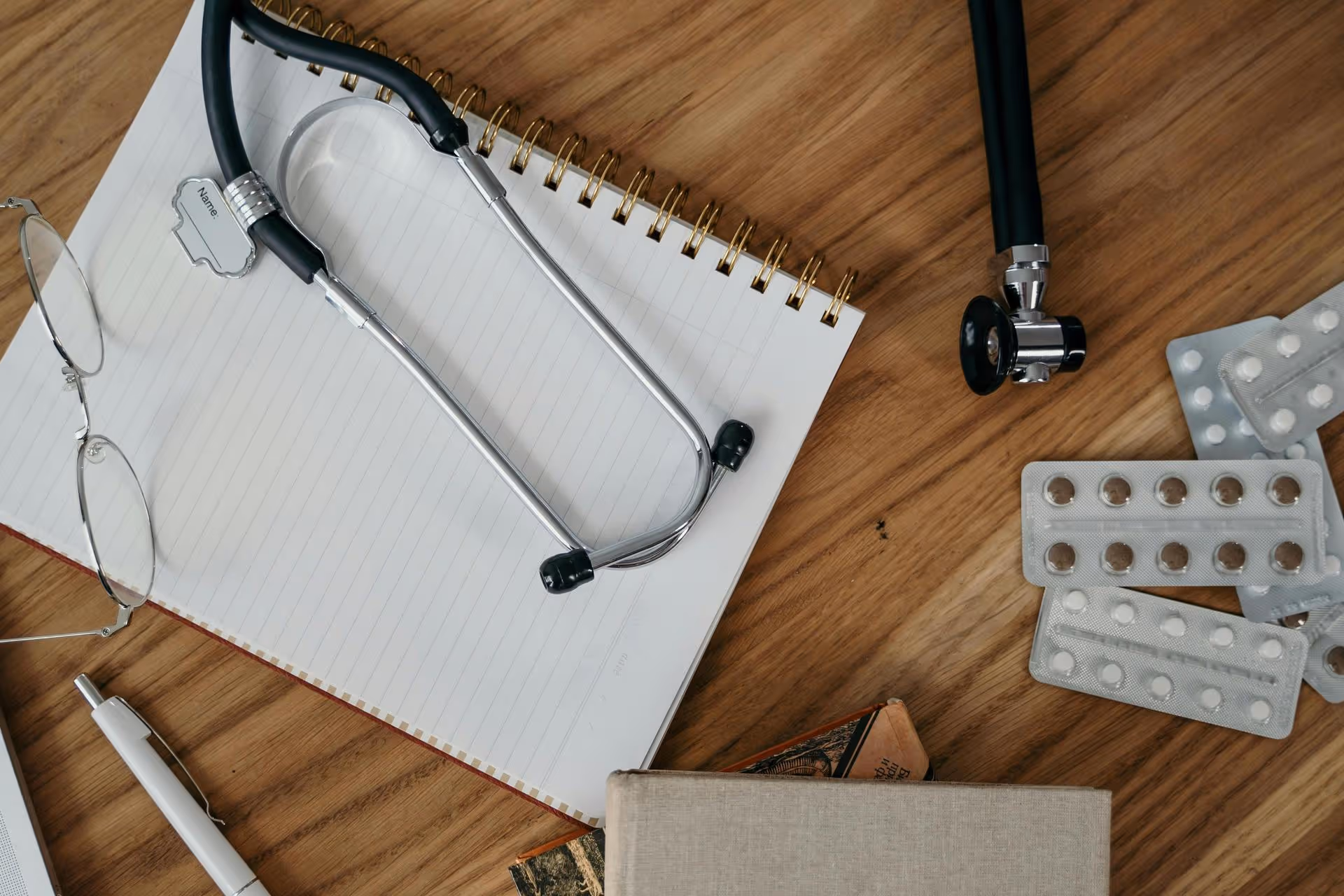 A stethoscope and glasses rest on a lined notebook on a wooden table, surrounded by scattered blister packs of pills, a pen, and a closed book, conveying a medical theme.