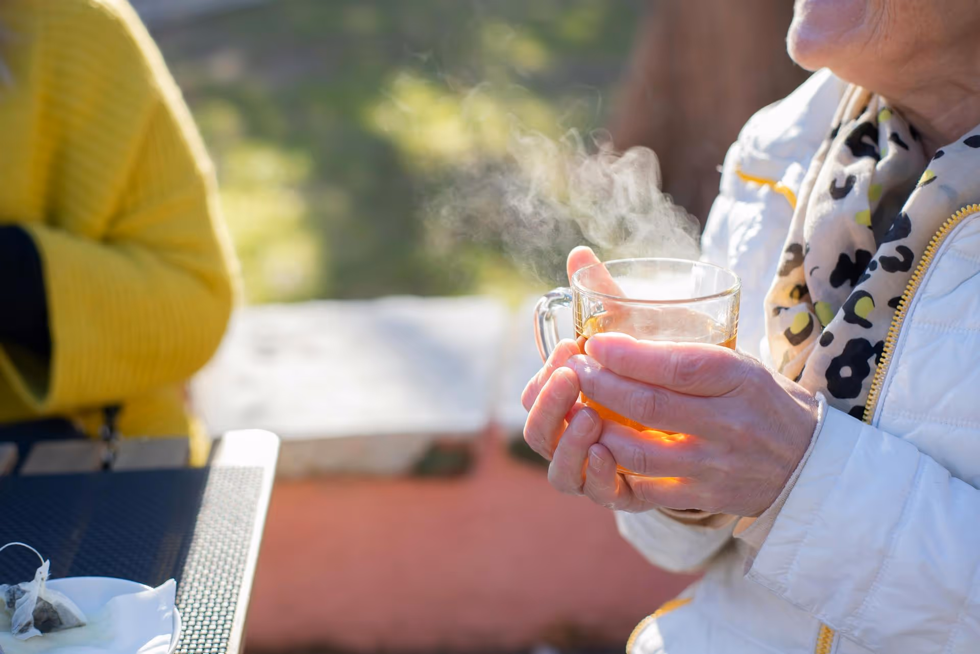 A person in a white coat holds a steaming glass of tea outdoors. The background is blurred, and another person in a yellow sweater is partially visible.