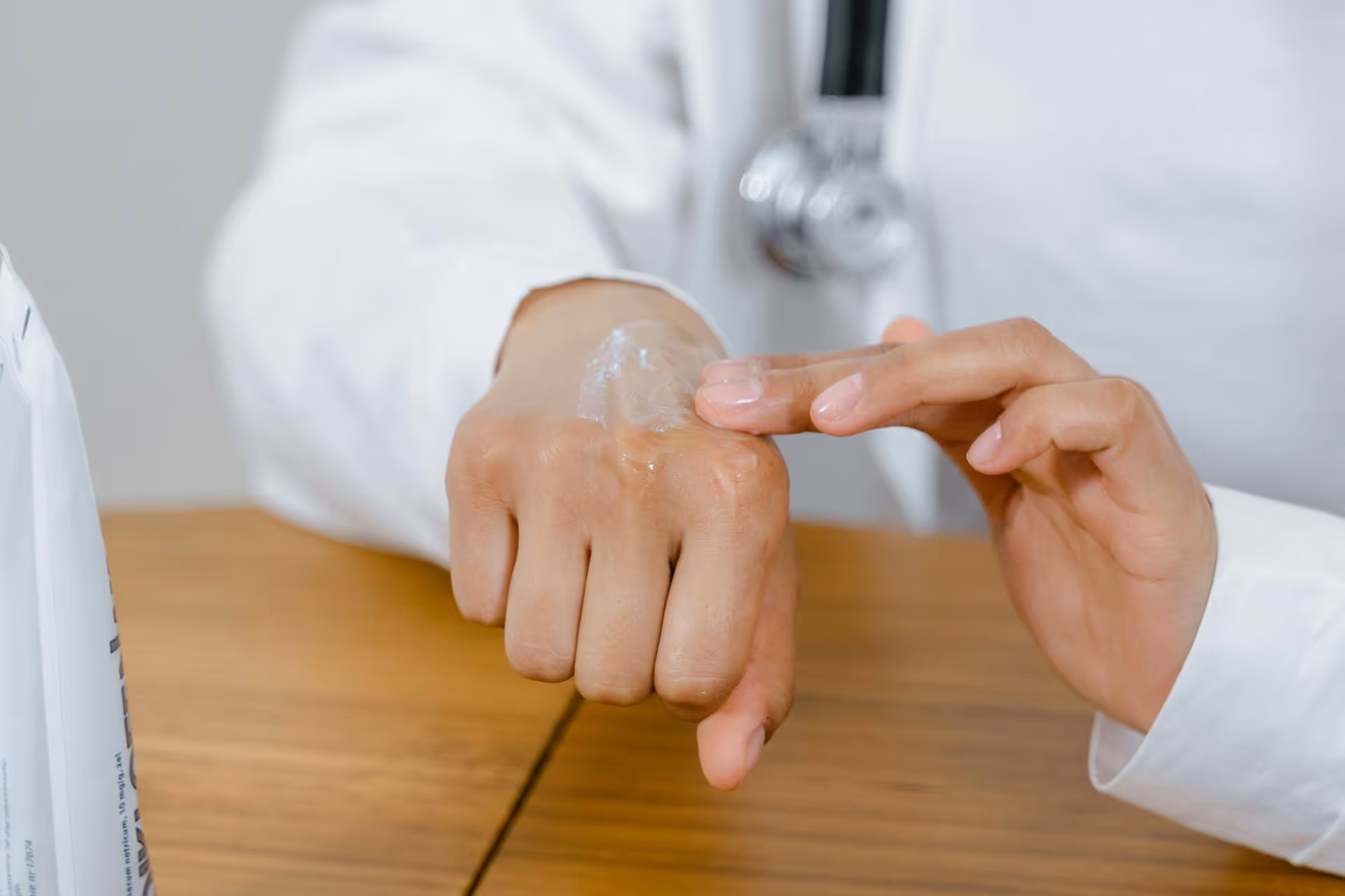 A person in a white coat applies a clear gel to the back of their hand, suggesting a medical or skincare setting. A stethoscope is partially visible.