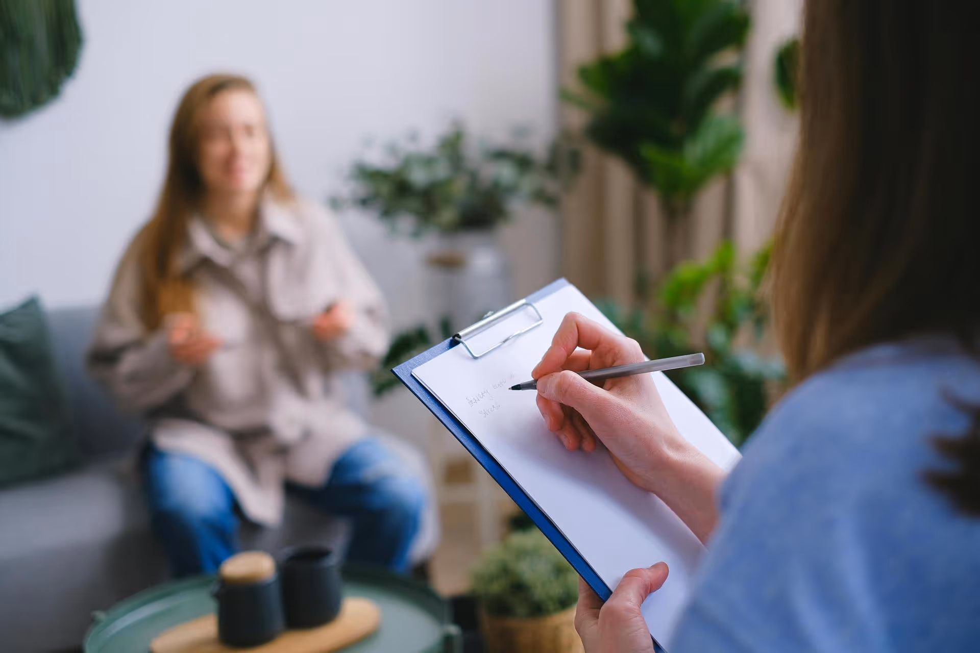 A therapist takes notes on a clipboard while listening to a woman sitting on a sofa. The room is calm, with soft lighting and green plants.