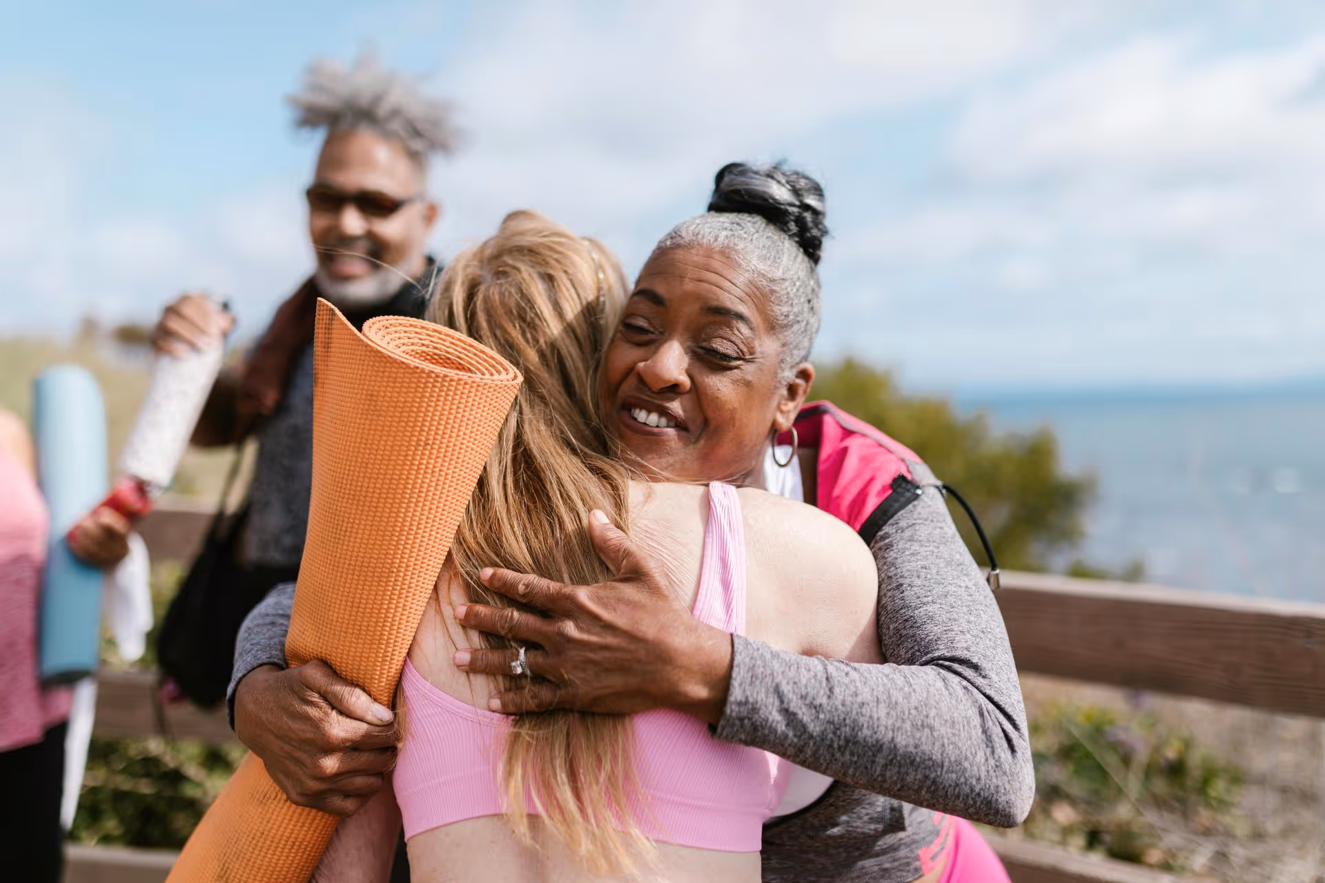 Two women warmly embrace, holding yoga mats, with a smiling man in the background. They are outdoors, conveying friendship and joy on a sunny day.