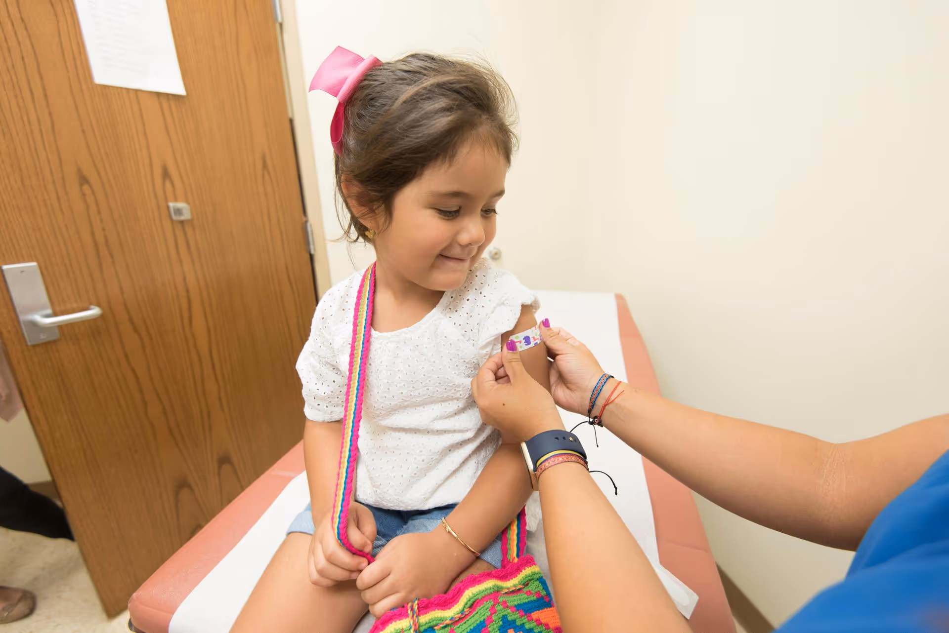 A young girl sitting on a doctor's examination table, smiling as a healthcare worker applies a colorful band-aid to her arm. She looks calm and content.