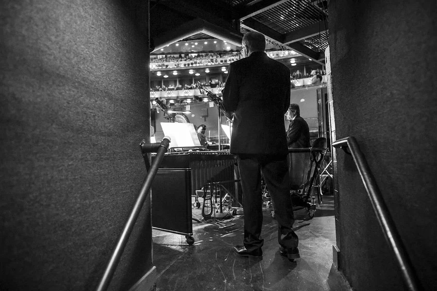 Black and white photo of musicians preparing backstage in a theater with audience seating visible in the background.