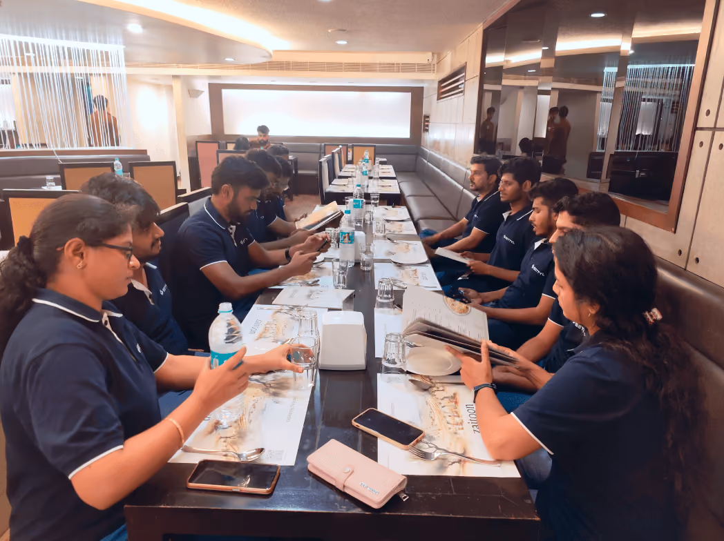 Group of people in matching navy blue shirts seated on both sides of a long dining table in a restaurant, looking at menus and smartphones.