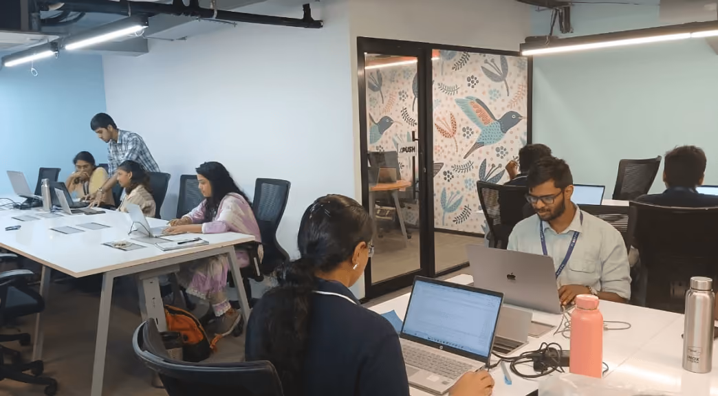 A modern office with several people working on laptops at white desks and a glass door featuring a decorative hummingbird mural.