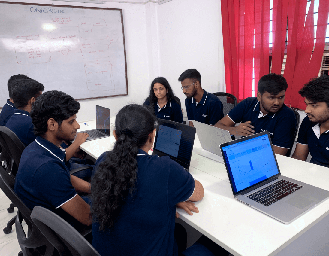 Group of seven people in navy blue polo shirts sitting around a white table with laptops, discussing in a meeting room with a whiteboard behind them.