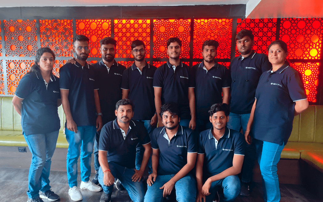 Group of eleven people wearing matching black SETTYL polo shirts posing indoors against a wall with decorative red and orange patterned panels.