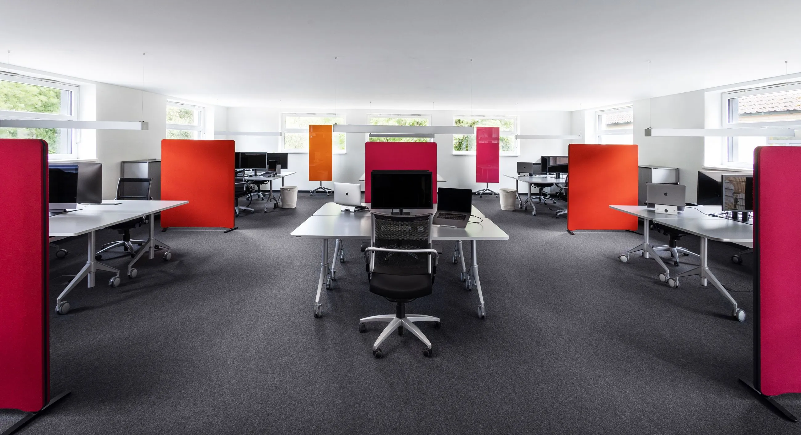 Modernes Büro mit mehreren weißen Schreibtischen, schwarzen Bürostühlen und farbigen Trennwänden in Rot und Orange.
