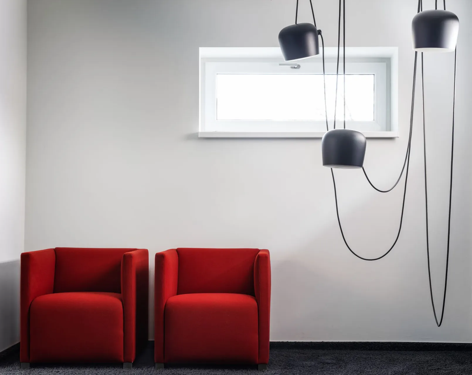 Zwei moderne rote Sessel nebeneinander vor einer hellen Wand mit kleinem Fenster und drei hängenden schwarzen Lampen.