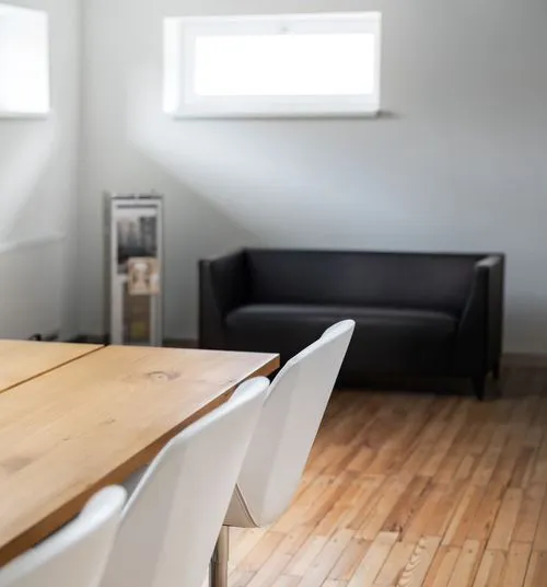 Moderner Besprechungsraum mit Holztisch, weißen Stühlen und schwarzem Sofa vor einer hellgrauen Wand mit kleinem Fenster.