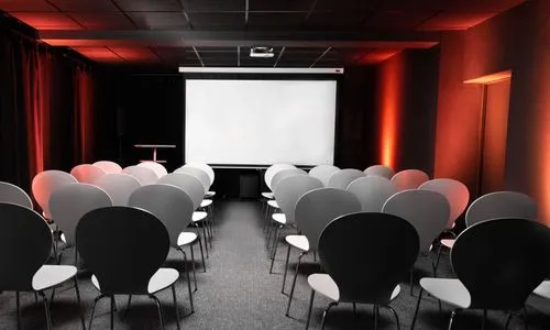Leerer Kinosaal mit Reihen von schwarzen und weißen Stühlen vor einer weißen Leinwand, von orangefarbenem Licht beleuchtet.