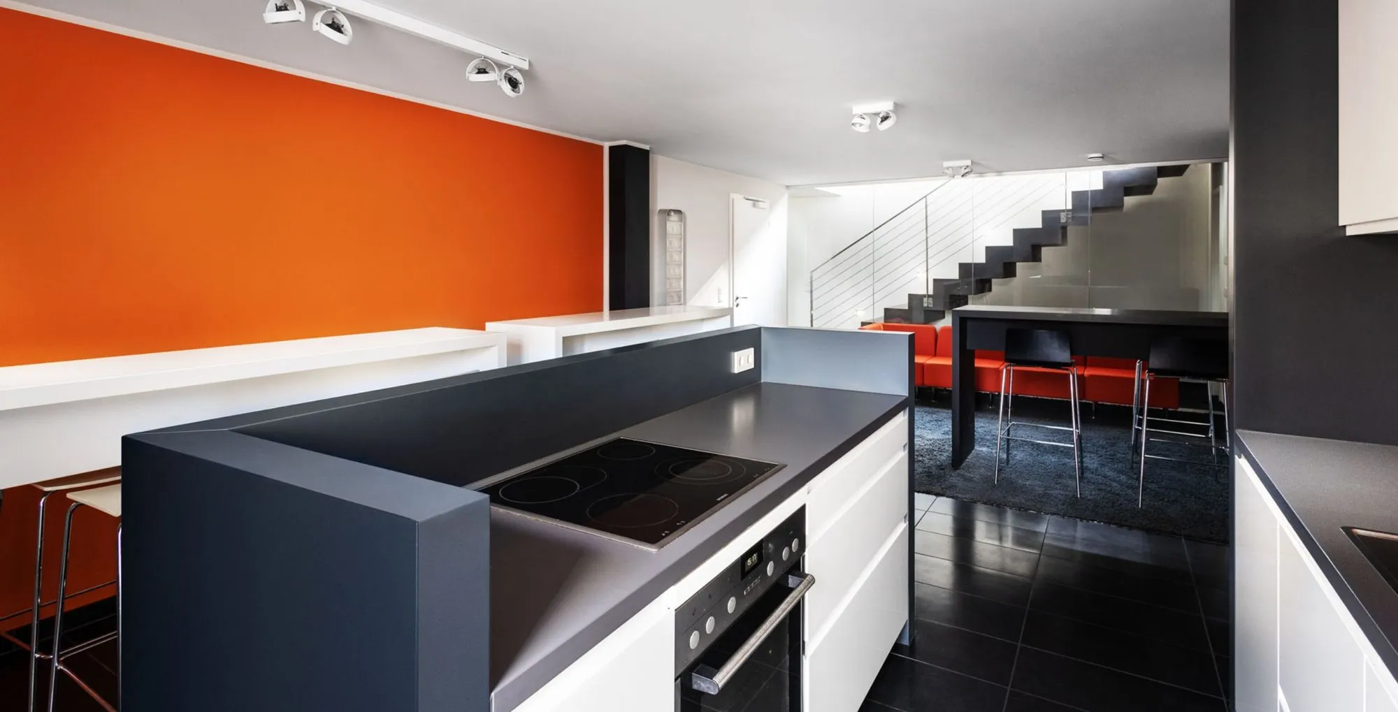 Moderne Küche mit schwarzer Kochinsel, weißen Schränken, oranger Wand und Wohnzimmer mit rotem Sofa und schwarzem Teppich.