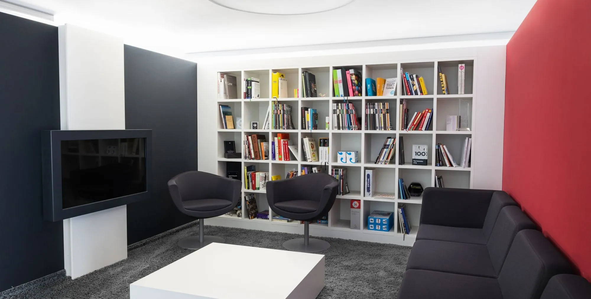 Moderner Wohnraum mit dunkelgrauem Sofa, zwei schwarzen Sesseln, weißem Couchtisch, großen Wandregalen mit Büchern und roter sowie schwarzer Wandfarbe.