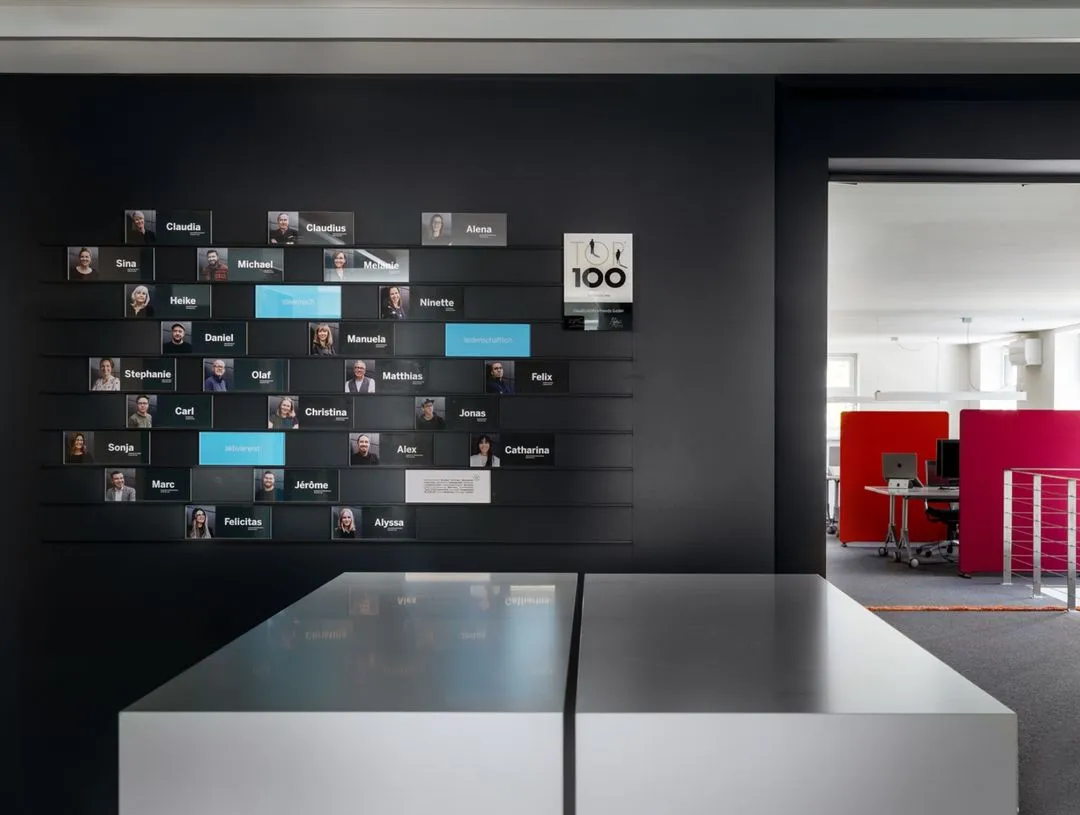Moderne Mitarbeiterwand mit Fotos und Namen vor schwarzer Wand in einem hellen Büro mit roten Raumteilern.