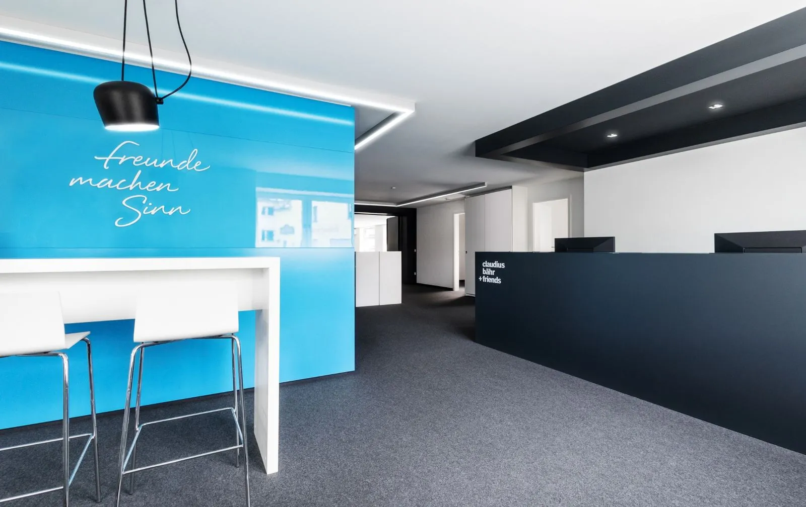 Moderner Empfangsbereich mit hellblauer Wand, zwei weißen Barhockern und dunklem Empfangstresen mit Monitoren.