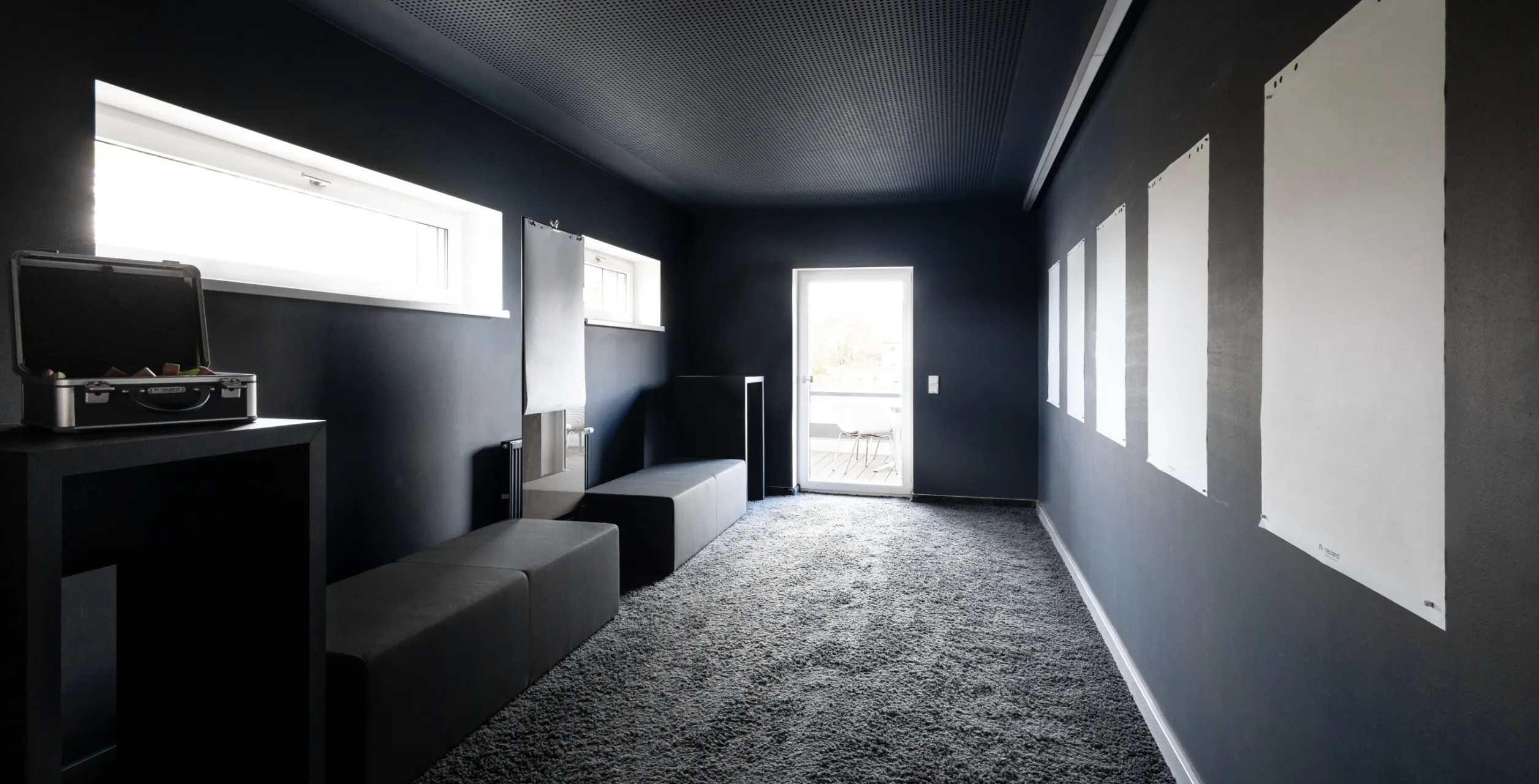 Dunkler Ruhe- oder Besprechungsraum mit grauem Teppich, schwarzen Sitzwürfeln, weißen Papierbögen an der Wand und einer hellen Tür zum Balkon.