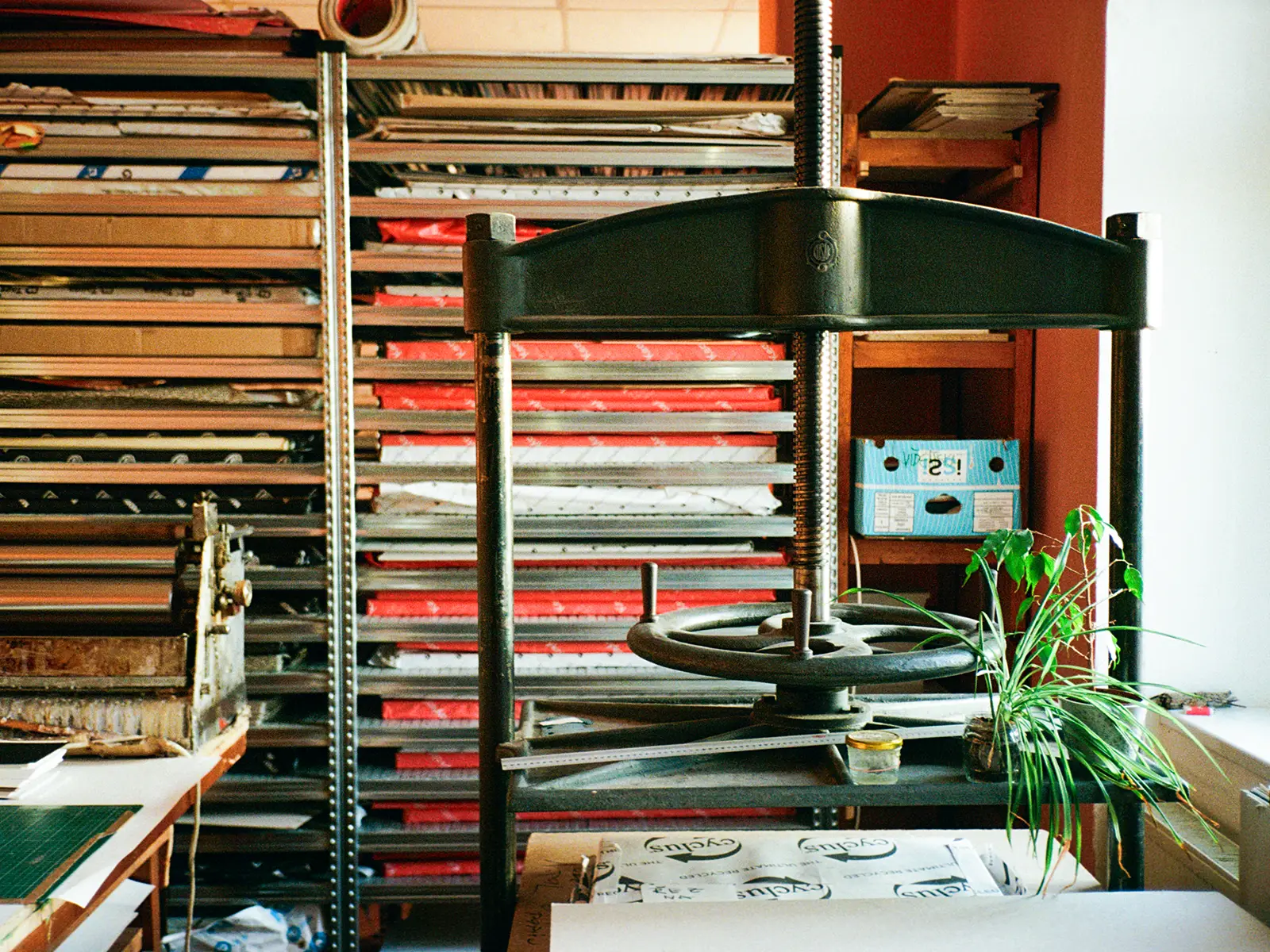 Ruční knihařský lis v tiskařském ateliéru, obklopený archivními regály s papírem, řezacím nástrojem a pracovním stolem s pravítky.