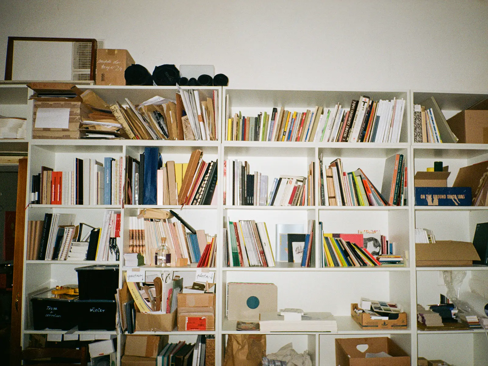 Ateliérový regál plný autorských knih, vzorkovníků, knihařských prototypů a tištěných publikací – dokumentace knižní tvorby a materiálů v kreativním studiu.