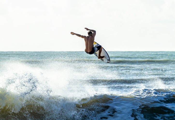 A surfer performs an aerial maneuver above a crashing wave, showcasing agility and skill. The ocean backdrop highlights a dynamic and adventurous scene.