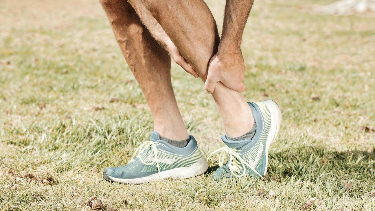 A person clutches their lower leg in apparent pain, wearing blue shorts and trainers. The setting appears to be outdoors on grass. The tone is one of discomfort.
