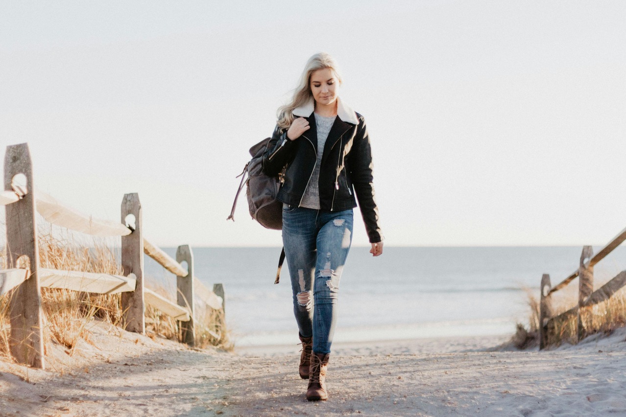 A woman walks down a sandy path toward the beach, carrying a backpack. She wears a black jacket and ripped jeans, evoking a calm and serene mood.