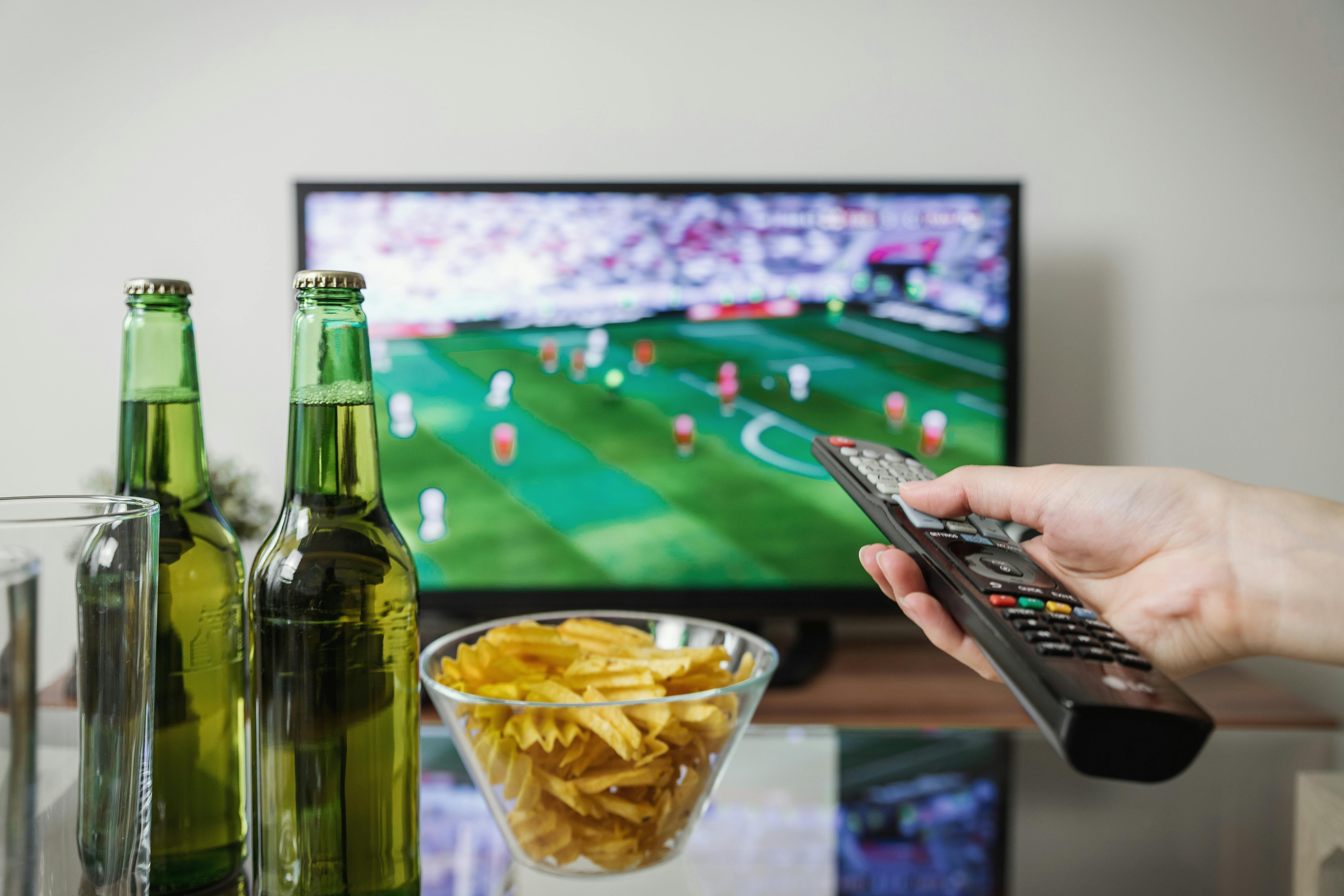 Person holding remote with football game on in the background and drinks and chips on the table