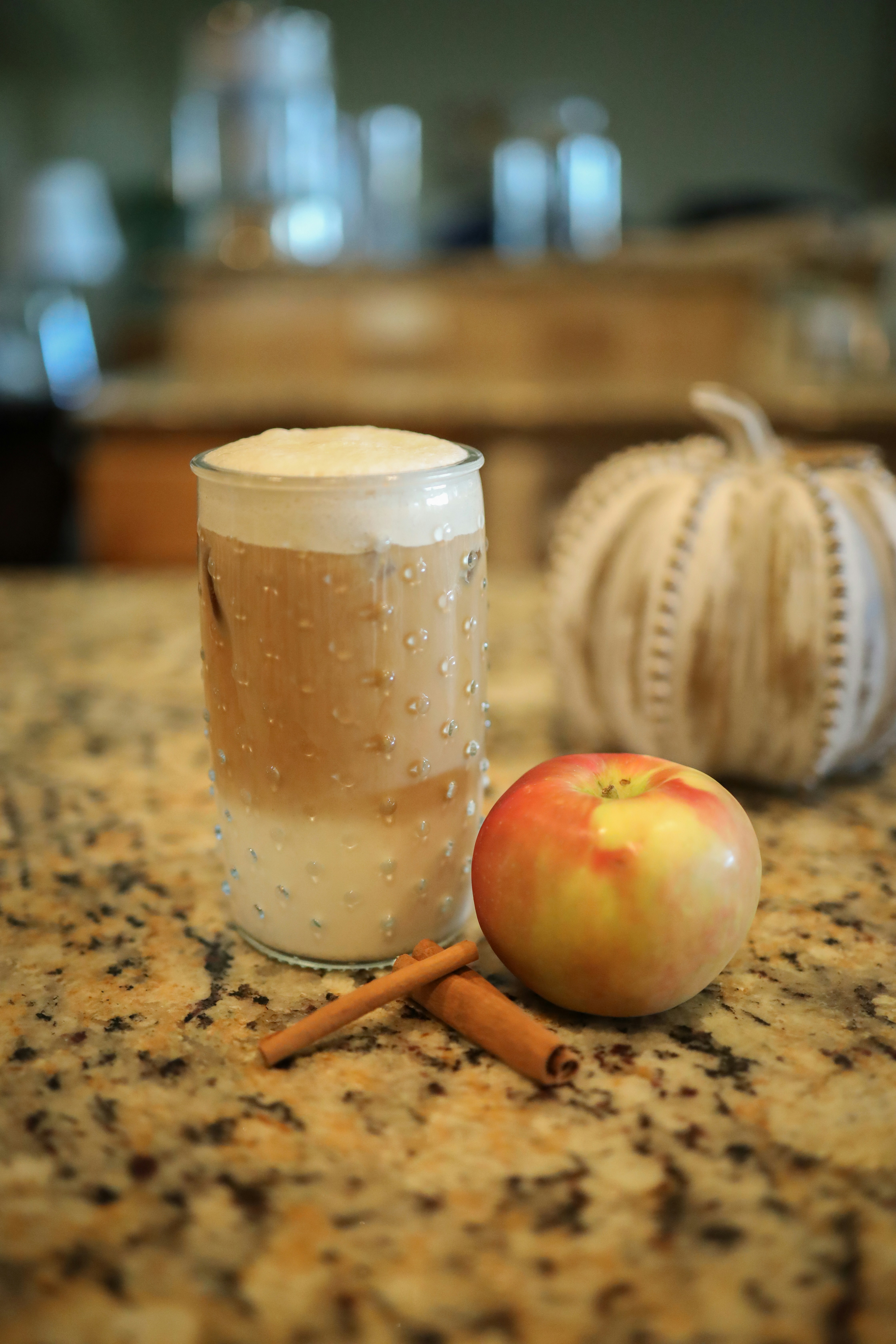 Glass of apple cider next to apple, cinnamon sticks, and decorative pumpkin on a kitchen counter