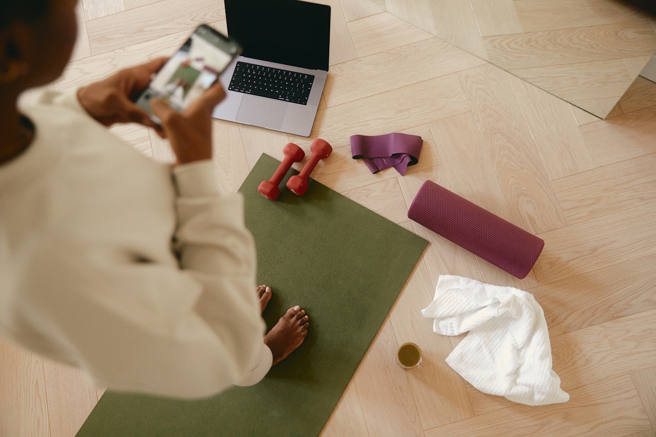 Young woman on phone in room with laptop, weights, yoga mat, and towel