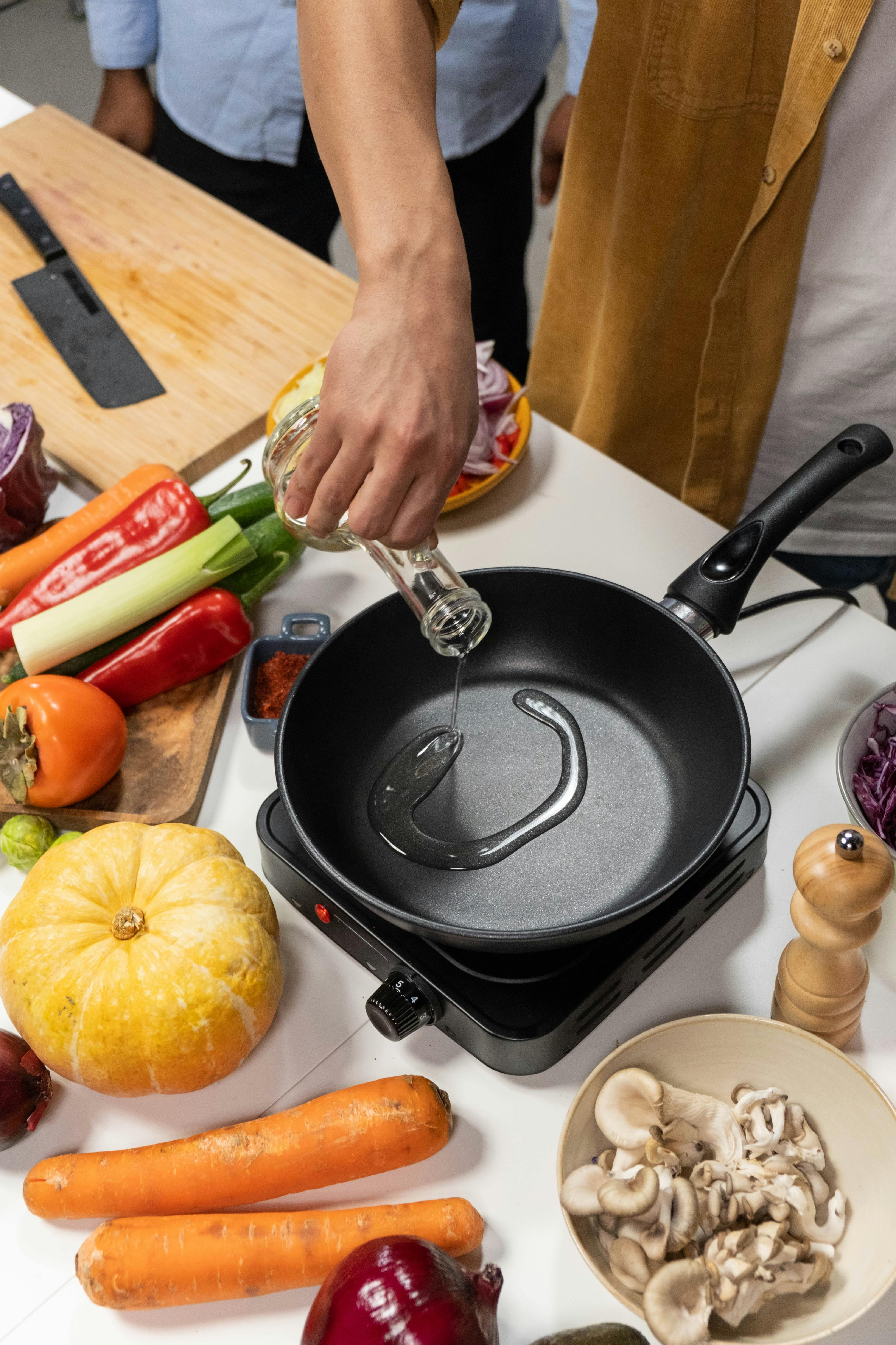 Person pouring oil into pan surrounded by vegetables.