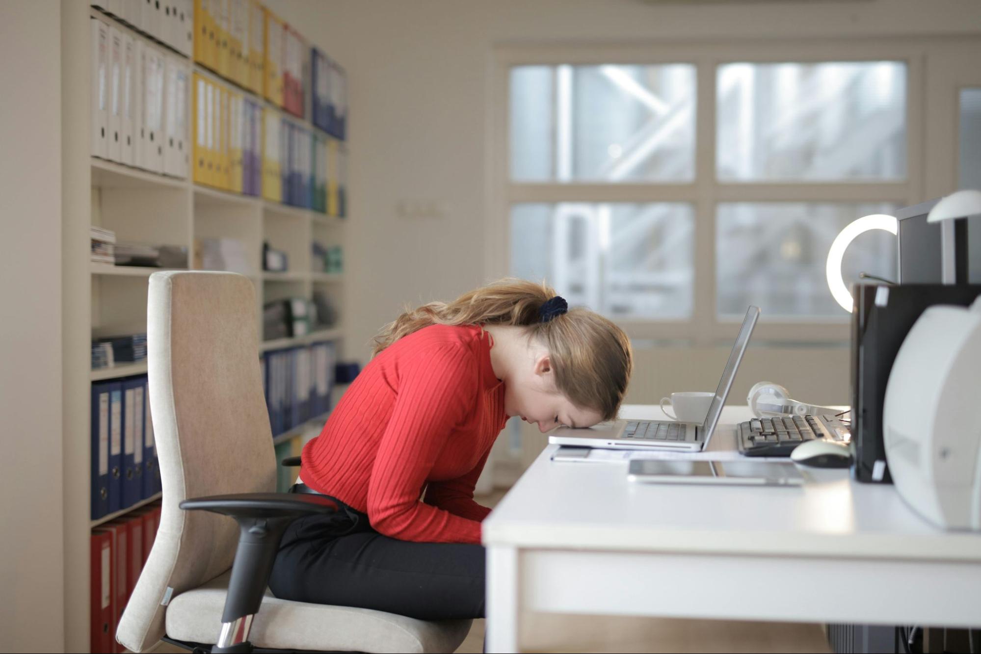 A tired woman in a red top is resting her head on her desk next to a laptop.