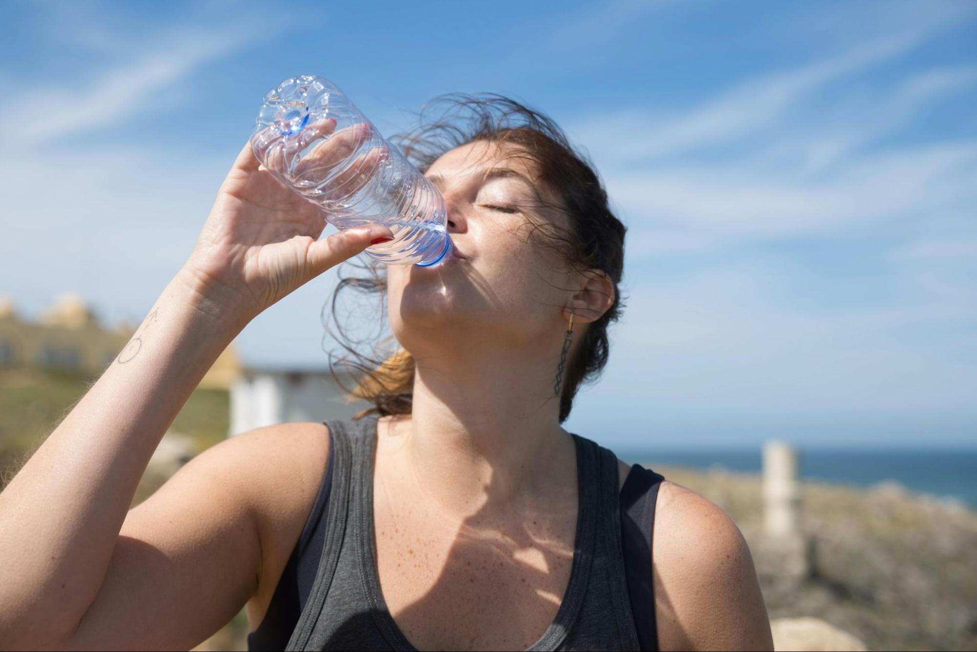 A woman in athletic wear is drinking water from a clear plastic bottle outdoors against a blue sky.