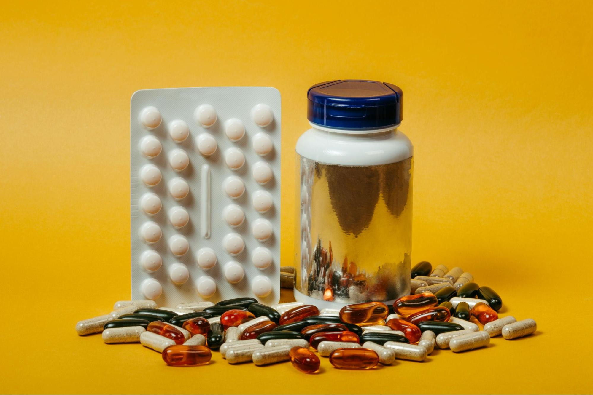 A pack of tablets, a supplement bottle, and scattered colorful capsules and pills arranged on a bright yellow background.