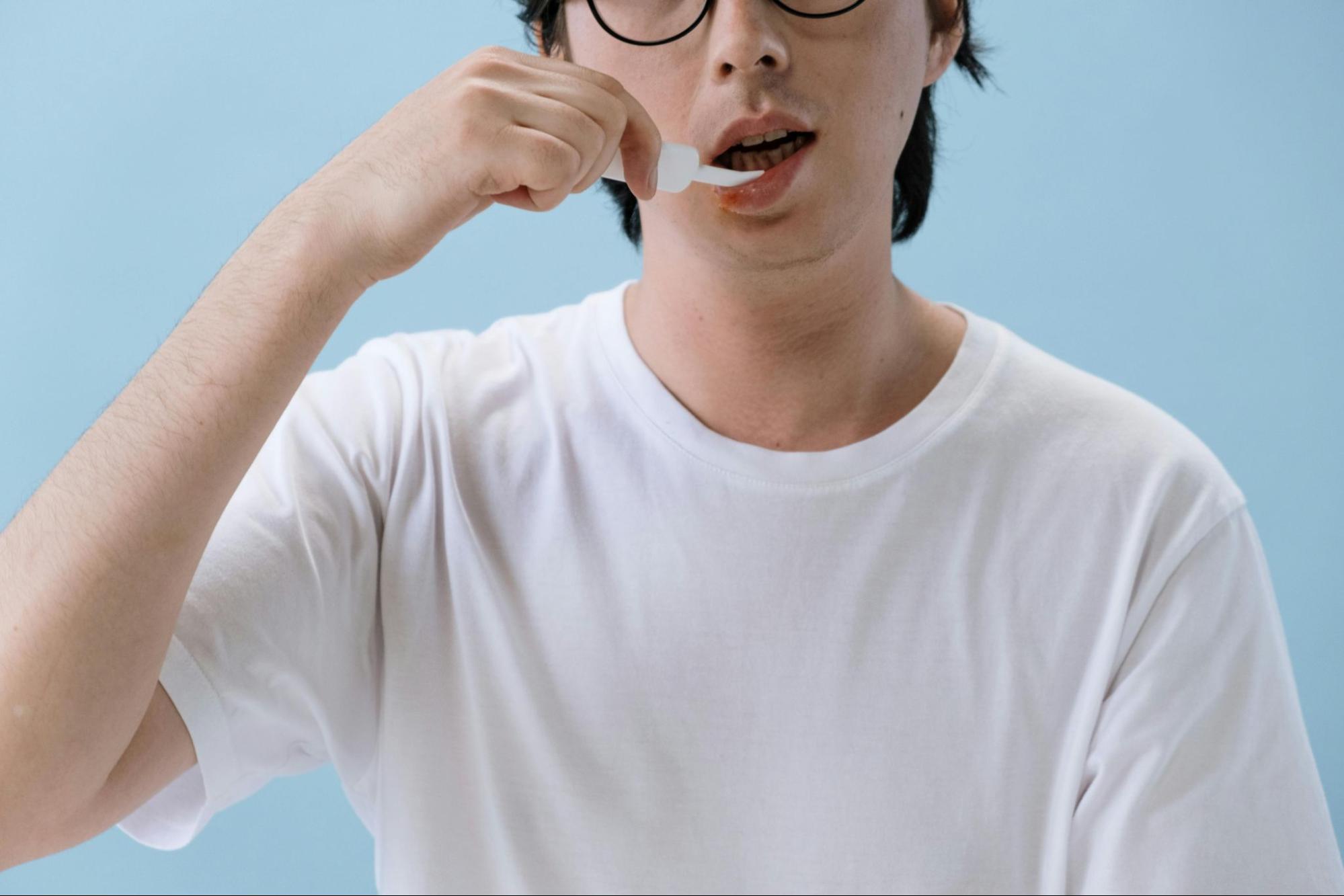 A person in a white t-shirt and glasses uses a dropper to place liquid medication under the tongue against a light blue background.