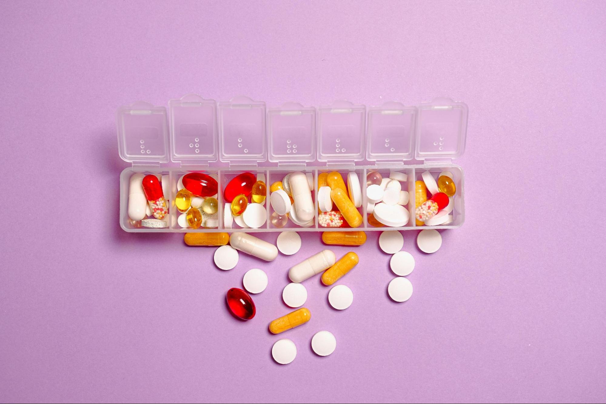 A weekly pill organizer with clear compartments contains various colorful vitamins and supplements on a pink background.