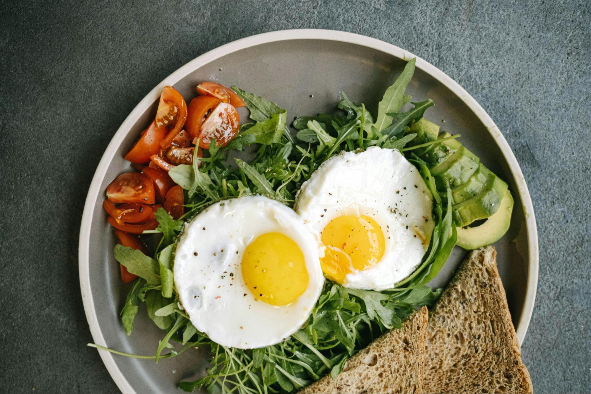 A gray plate with over-easy eggs on a bed of arugula, toast, and sliced tomato and avocado.