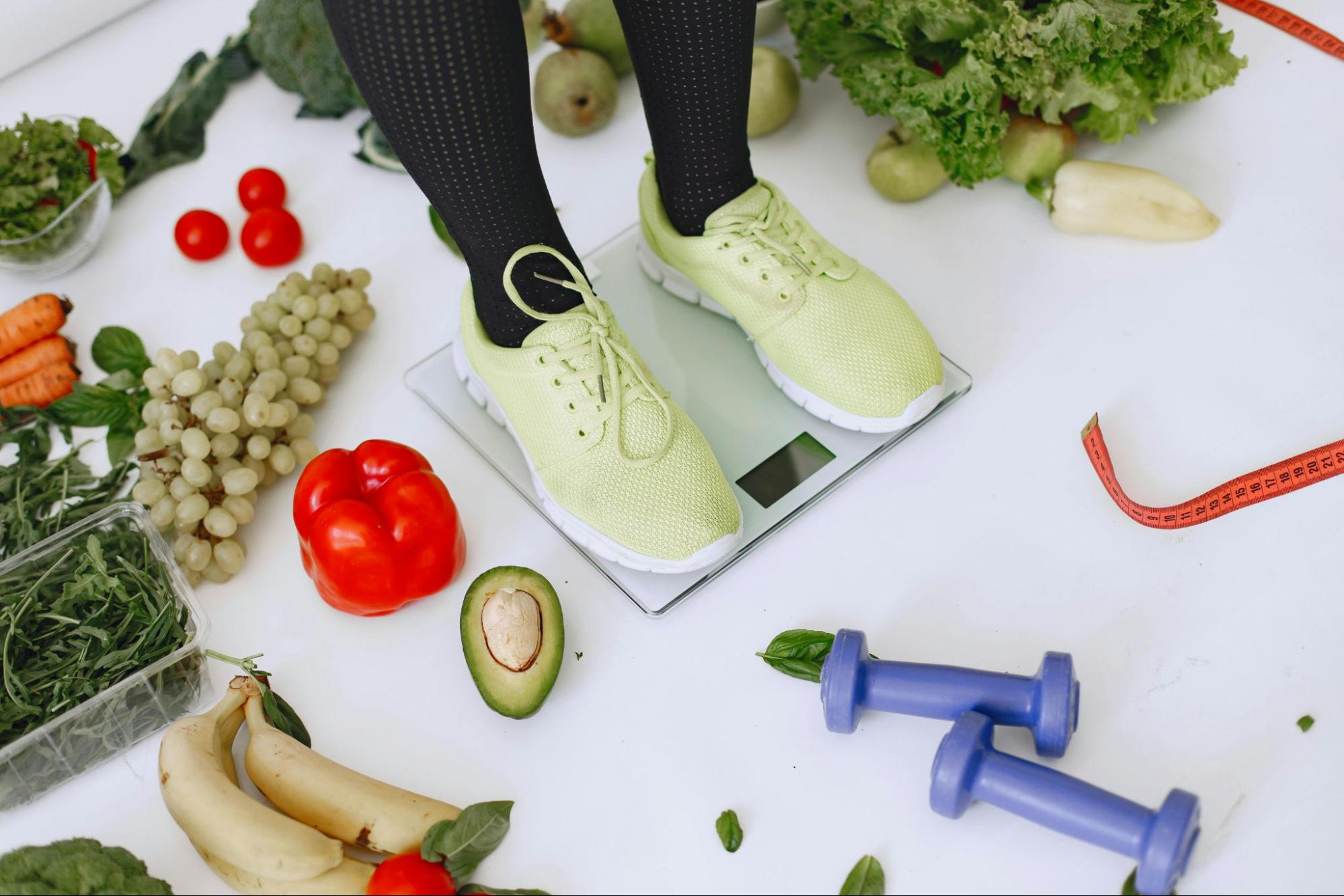 Person standing on a weight scale surrounded by fresh vegetables, fruit, and dumbbells.